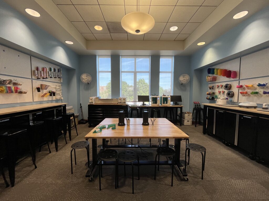 interior of the Cranium makerspace with sunlight and room lights on, clear table surrounded by work stools, yarn and other equipment on the walls, and 3D printers and other equipment on tables along the walls